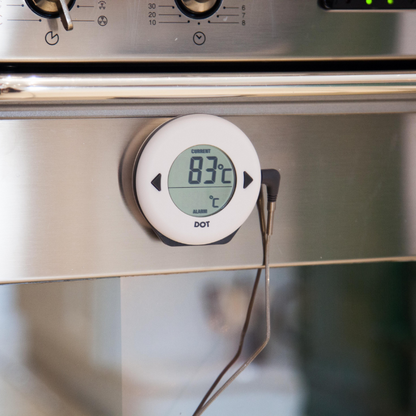 Een witte, ronde digitale thermometer met zwarte knoppen en een helder scherm dat 34°C huidtemperatuur en 75°C alarm toont. Geschikt voor medische of persoonlijke temperatuurcontrole.