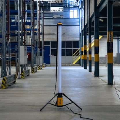 Een witte, opstelbare werklicht met gele basis en zwarte driepoot, voorzien van een kabel en schakelaar, geschikt voor gebruik in werkplaatsen en bouwprojecten.