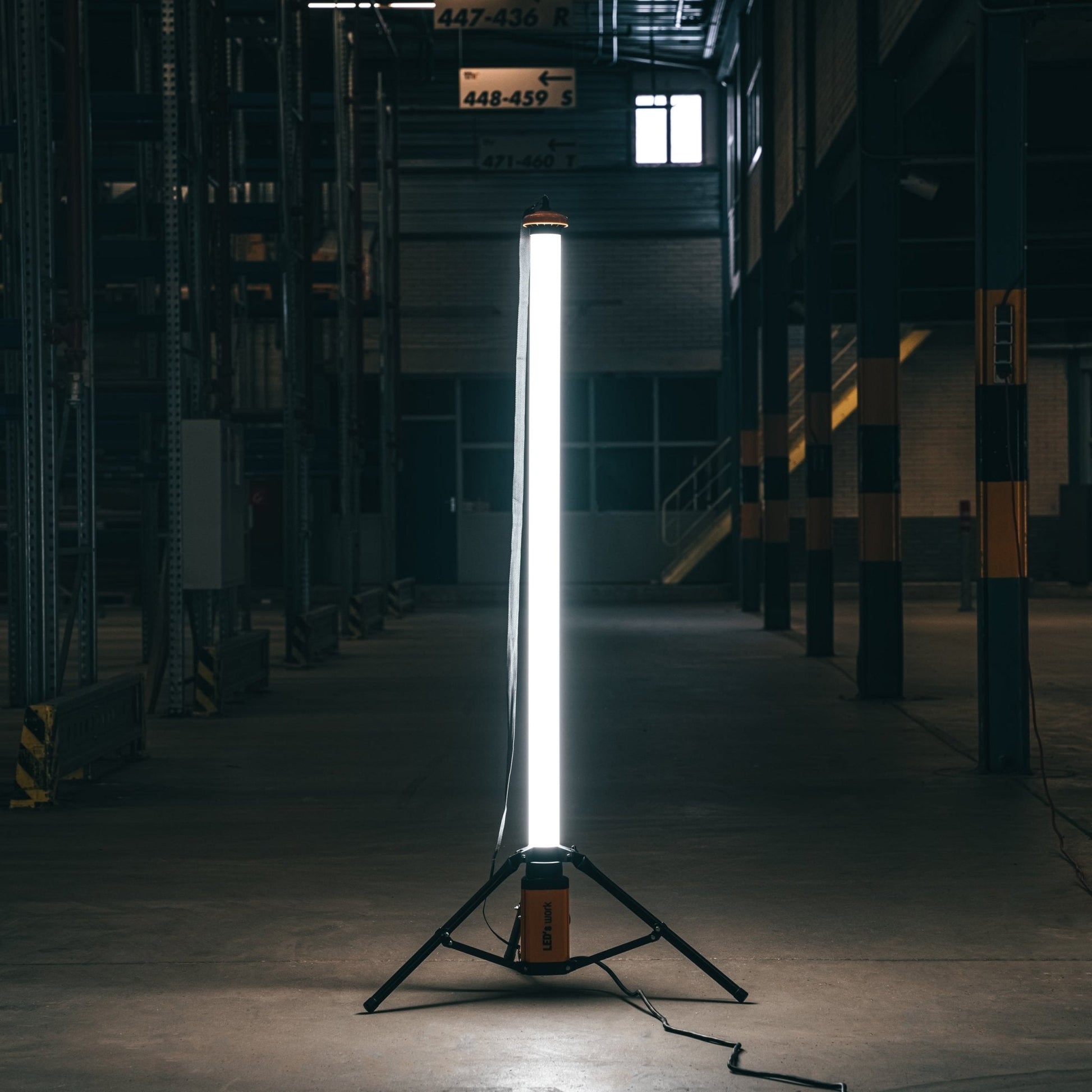 Een witte, opstelbare werklicht met gele basis en zwarte driepoot, voorzien van een kabel en schakelaar, geschikt voor industriële of bouwplaatsverlichting.