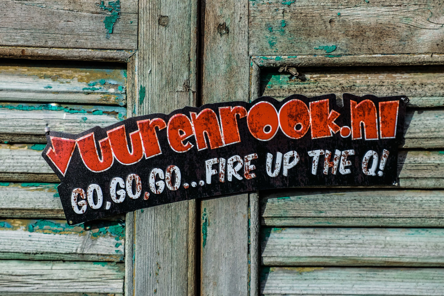 Een roestig zwart metaalplaatje met rode en witte letters, waarop ‘vuurenrook.nl’ en ‘GO,GO,GO...FIRE UP THE Q!’ staat, bedoeld als decoratief element voor barbecue- of grillfeesten.